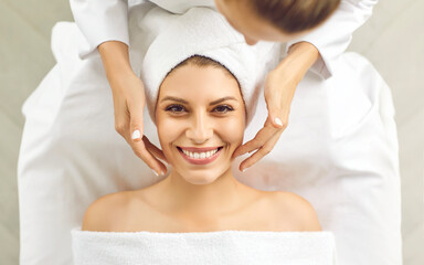 Wall Mural - Cheerful young woman enjoying facial treatment at spa salon or beauty parlor. Top overhead view from above close up portrait happy joyful woman in white towel turban on bed. Skin care, beauty concept