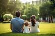 © tohceenilas - A family of three sits on green grass in a park, enjoying a sunny day surrounded by trees and nature, creating a serene and peaceful moment.