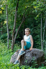 Wall Mural - Portrait of a lively cute boy sitting on a stone on a beautiful green background of trees