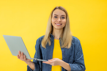 cheerful young woman standing isolated over yellow background using laptop computer. Looking at camera.