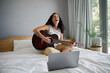 © Nicola Suttle/Stocksy - Cheerful woman playing guitar on bed with laptop