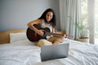 © Nicola Suttle/Stocksy - Happy young woman learning playing guitar on bed