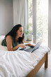 © Nicola Suttle/Stocksy - Smiling woman browsing computer on soft bed