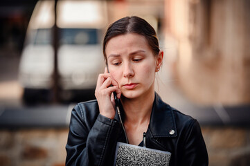 Poster - A young woman in a black leather jacket holds a notebook and talks on her phone, appearing focused and serious in an urban setting.