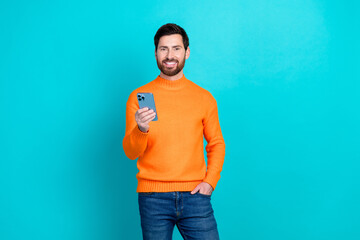 Poster - Photo of cheerful handsome man wear orange stylosh clothes hold phone isolated on cyan color background