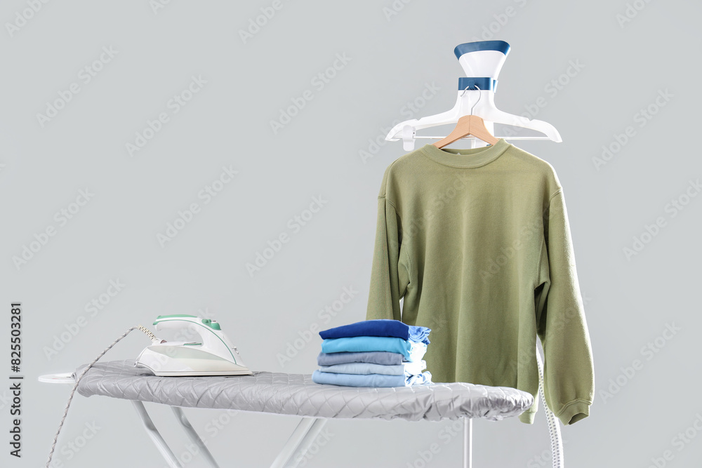 Ironing board with stack of clean clothes and garment steamer on grey background