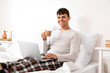 © Pixel-Shot - Young man with cup of coffee using laptop in bedroom