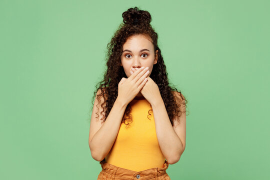 Young sad shocked surprised woman of African American ethnicity wear yellow tank shirt top look camera cover mouth with hands isolated on plain pastel light green background studio. Lifestyle concept.