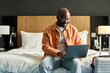 © Mediaphotos - Portrait of smiling Black man using laptop sitting on bed in hotel room and researching accommodation facilities copy space