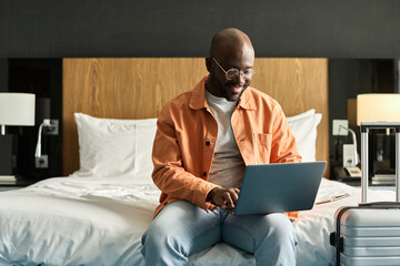 Portrait of smiling Black man using laptop sitting on bed in hotel room and researching accommodation facilities copy space