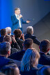© Anton Gvozdikov - Speaker is addressing an audience at a business conference. People in the audience are paying attention and some are taking photos.