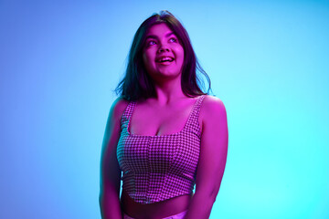 Young Indian girl, 20s years old, looking away with playful expression in pink neon light against gradient blue background. Concept of human emotions, beauty and fashion, youth. Ad