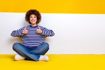 Full length photo of cool woman dressed striped shirt sit near placard empty space show thumbs up isolated on yellow color background