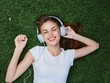 © SHOTPRIME STUDIO - Woman listening to music with headphones in a white T-shirt on the green lawn grass in the park, vacation smile with teeth, looking into the camera