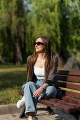 Attractive caucasian young woman in sunglasses and stylish casual clothes sitting on a park bench on a warm spring day. Rest, freedom, leisure.