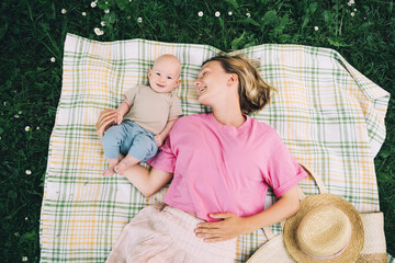 Wall Mural - Smiling mom and baby lying on blanket on green grass at summer. Family relaxing and having picnic outdoors. Beautiful mother with her baby on nature. Concept of motherhood, human happiness, eco life.