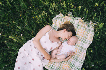 Wall Mural - Smiling mom and baby lying on blanket on green grass at summer. Family relaxing and having picnic outdoors. Beautiful mother with her baby on nature. Concept of motherhood, human happiness, eco life.