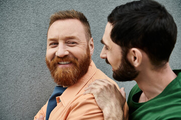 A gay couple in casual attire stand side by side against a grey wall, showing their love for each other.