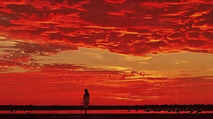 Wall Mural -   Person on beach with surfboard beneath red-yellow sky, clouds in background