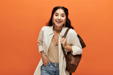 Smiling indian woman with backpack against vibrant orange backdrop.