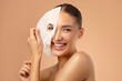 © Home-stock - Young pretty European woman removing moisturizing, rejuvenating white fabric sheet mask from her face after using it, smiling at camera, beige background