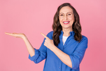 Sticker - Photo portrait of attractive retired woman point hold empty space dressed stylish blue clothes isolated on pink color background