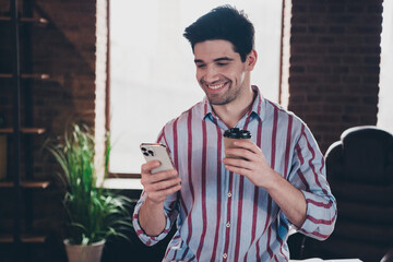 Wall Mural - Photo of young man corporate worker use smart phone drink coffee wear striped shirt modern office home indoors