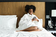 © Mediaphotos - Portrait of young Black woman wearing bathrobe and using smartphone while relaxing on bed in hotel room, copy space