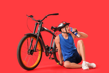 Wall Mural - Young man sitting near bicycle and drinking water on red background