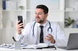 © New Africa - Doctor having online consultation via smartphone at table in clinic
