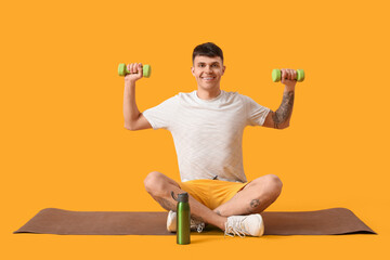 Wall Mural - Sporty young man with bottle of water and dumbbells sitting on mat against yellow background