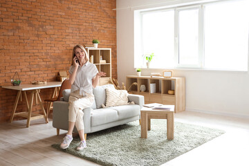 Sticker - Young pretty woman sitting on sofa and talking with mobile phone on sofa in living room