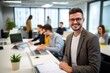 © alisaaa - Portrait of a young male IT professional sitting at desk, holding document, smiling and looking at camera at office with his coworkers in background