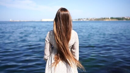 Wall Mural - A woman with long brown hair stands on a beach, looking out at the ocean. The water is calm and the sky is clear, creating a peaceful and serene atmosphere.
