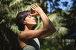 © insta_photos - Calm young Hispanic woman holding hands in namaste meditating doing yoga breathing exercises with eyes closed feeling peace of mind, mental balance standing in green nature tropical park.