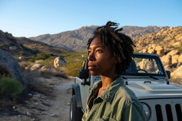 Wall Mural - A woman with dreadlocks stands in front of a jeep. The jeep is parked on a dirt road in the desert. The woman is looking at something in the distance