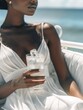 © vefimov - A woman in a white dress holding a glass of water with ice cubes in it. Concept of relaxation and leisure, as the woman is sitting on a couch by the ocean