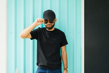 Hispanic man exudes confidence in a black t-shirt and cap against a turquoise backdrop, ideal for mockup designs