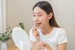 © KMPZZZ - Lip care concept, happy asian young woman finger touching lips in front of mirror at home after applying lipstick balm on dry mouth from natural beauty product, skincare routine, makeup and cosmetics.