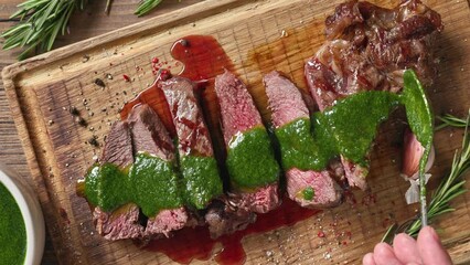 Poster - Chef pours green parsley sauce on a slices of freshly grilled beef steak meat on a wooden cutting board. Close-up of food, top view