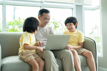 Asian father and his two sons have fun watching video in laptop while sitting on sofa in living room at home. Family relaxing on holiday.