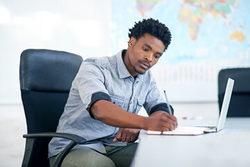 Desk, thinking and black man with laptop, notes or business plan at international agency. Research, consultant or travel agent in office writing info in notebook for schedule agenda or online booking