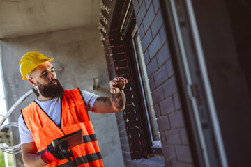 Portrait of successful man constructor wearing yellow helmet and safety yellow vest. Portrait of architect standing at building site and using tablet. He is inspecting how the work is going.