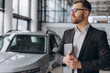 © anatoliycherkas - Modern bearded man in glasses and suit vehicle sales consultant using laptop inside car dealership
