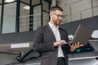 © anatoliycherkas - Modern bearded man in glasses and suit vehicle sales consultant using laptop inside car dealership