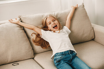 Cheerful young girl with arms raised, sitting on a cozy couch, expressing joy and happiness in a bright living room