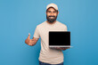 © Prostock-studio - Indian man is standing while holding a laptop with black screen in one hand and giving a thumbs up gesture with the other hand
