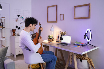 Wall Mural - Male student playing guitar while streaming at home in evening