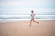 © Andrii - Full length of athletic woman running along seashre. Side view of young sportswoman in white top and sporty shorts jogging on sandy beach. Fit lady represents healthy lifestyle.
