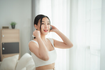 Wall Mural - Joyful young woman with long dark hair, wearing a white crop top and beige pants, smiling brightly as she adjusts her headphones. She is in a bright, modern living room, exuding happiness and energy.